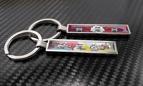 Porte-clés style bande dessinée avec plaque d’immatriculation et illustrations de voitures, placés sur une surface à texture carbone